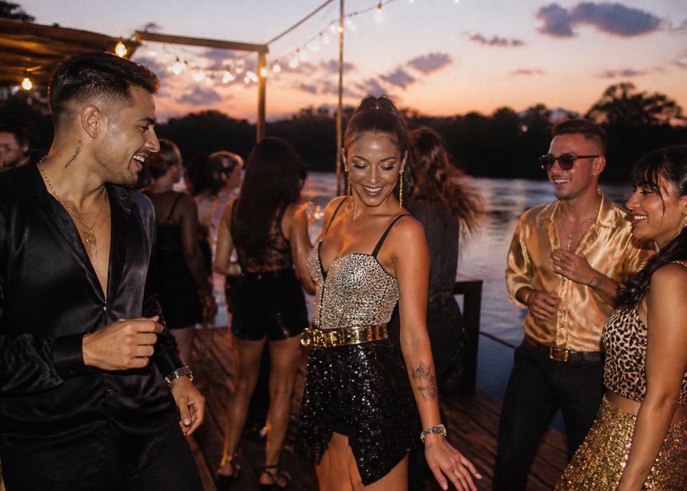 Invitados bailando en el muelle al atardecer con looks de brillo y satén